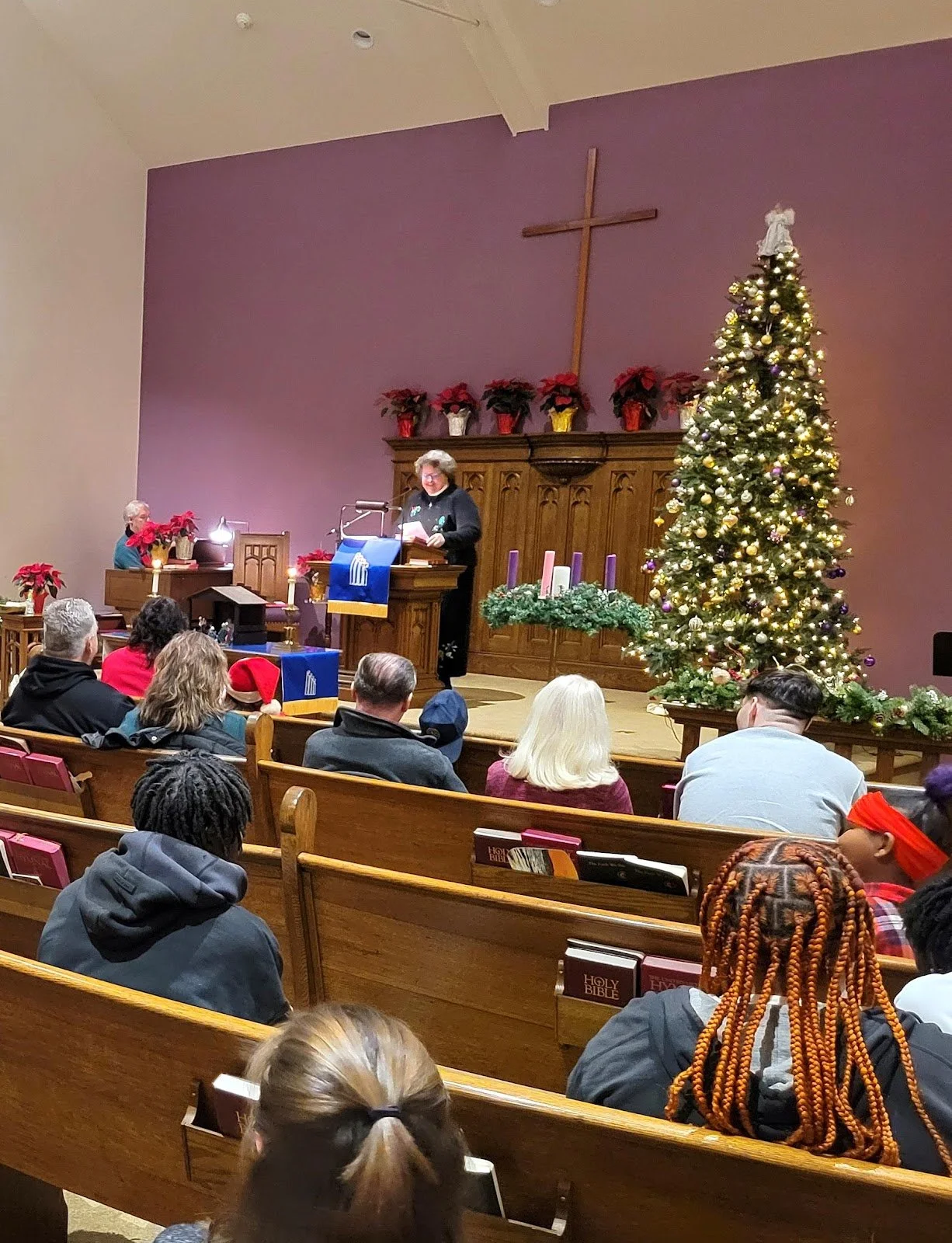 Church interior decorated for Christmas with a Christmas tree, poinsettias, candles, and a cross on the wall, with a speaker at the pulpit and congregation seated.