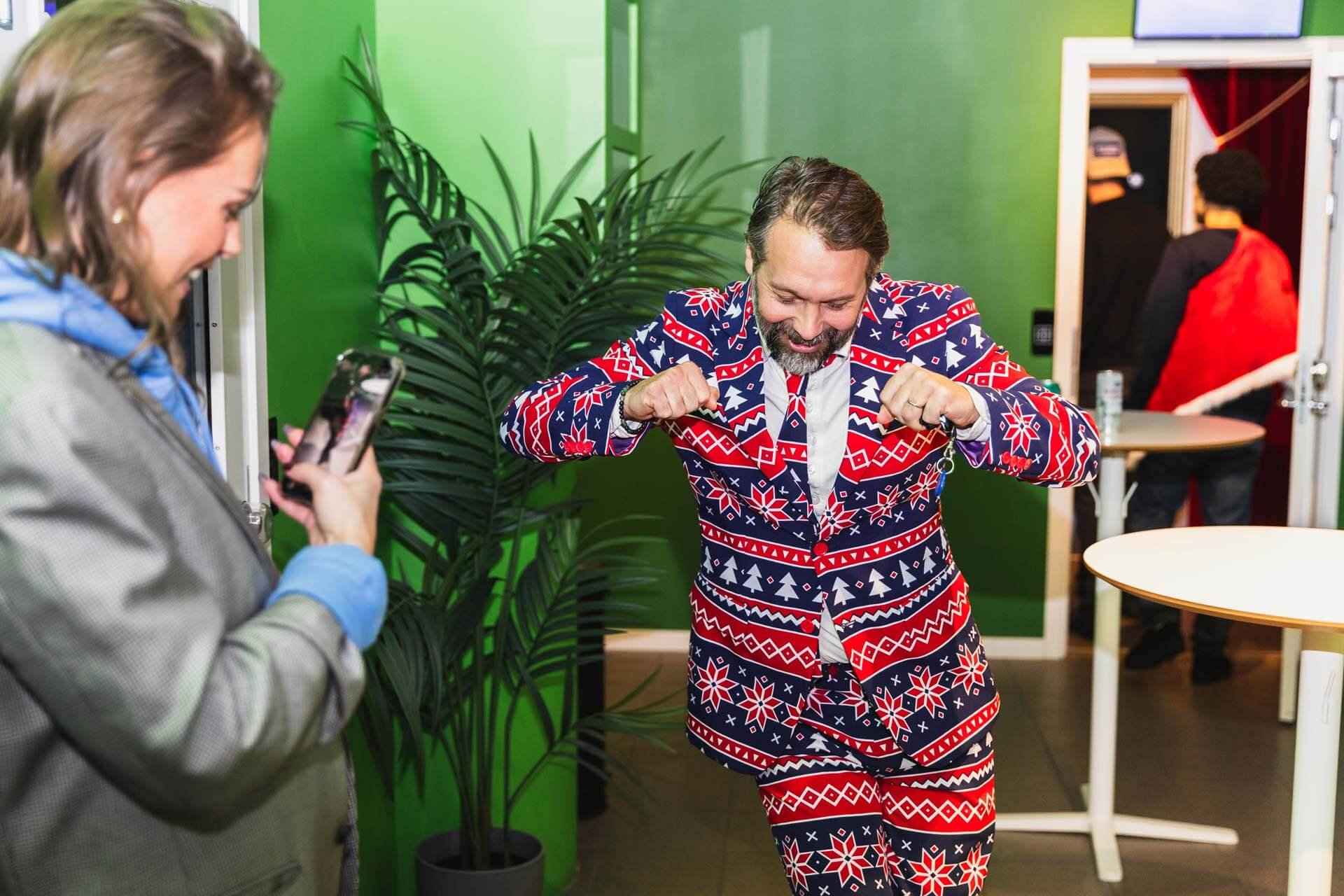 Man i julig inrättning, klädd i en julig kostym, visar musklerna medan en kvinna tar ett foto av honom. I bakgrunden en man i svart och röd jacka.