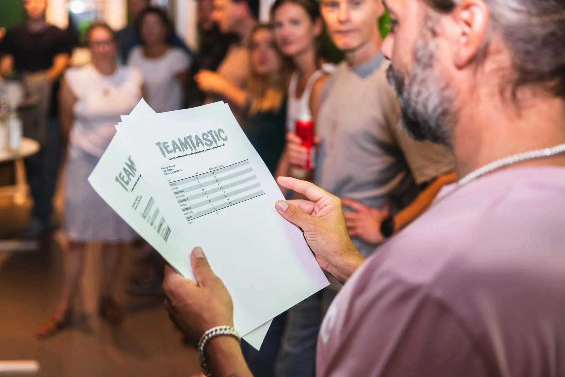 En man håller i två papper med rubriken 'TEAMSTASTIC' medan en grupp människor står i bakgrunden, troligen i en social eller sportrelaterad miljö.