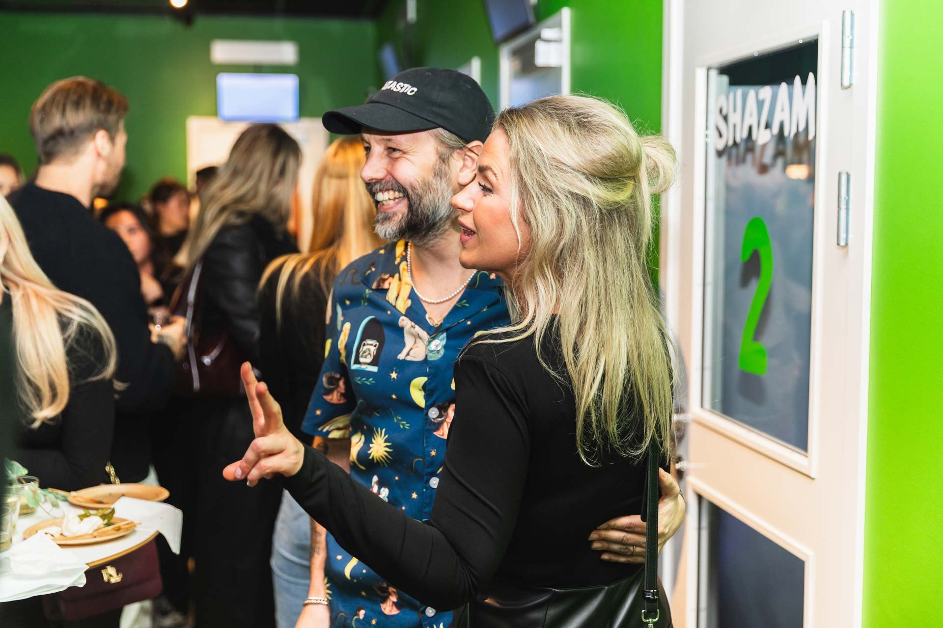 Två personer säger hej och ler under en social sammankomst i ett rum med gröna väggar.