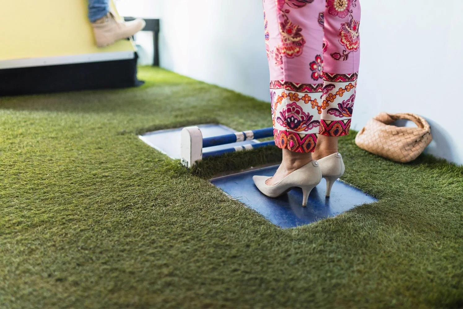 En person i färgglada byxor med höga klackar står på ett litet blått plattform, troligen ett steg eller en låg bro, som är placerat på gräsmatta. I bakgrunden finns ett annat ben av en person med ljus träskor och en svart möbel.