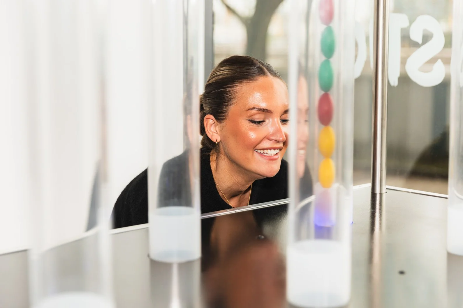 En kvinna med lockigt hår och svart tröja som ler när hon tittar på ett experiment med vätskelabbar i ett laboratorium.