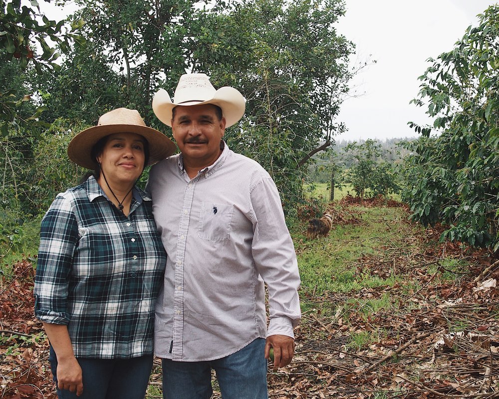 A man and woman standing outdoors in a green, tree-filled area, both wearing hats and smiling at the camera. Armando and Karina, founders of Aloha Star 100% Kona Coffee Farm.