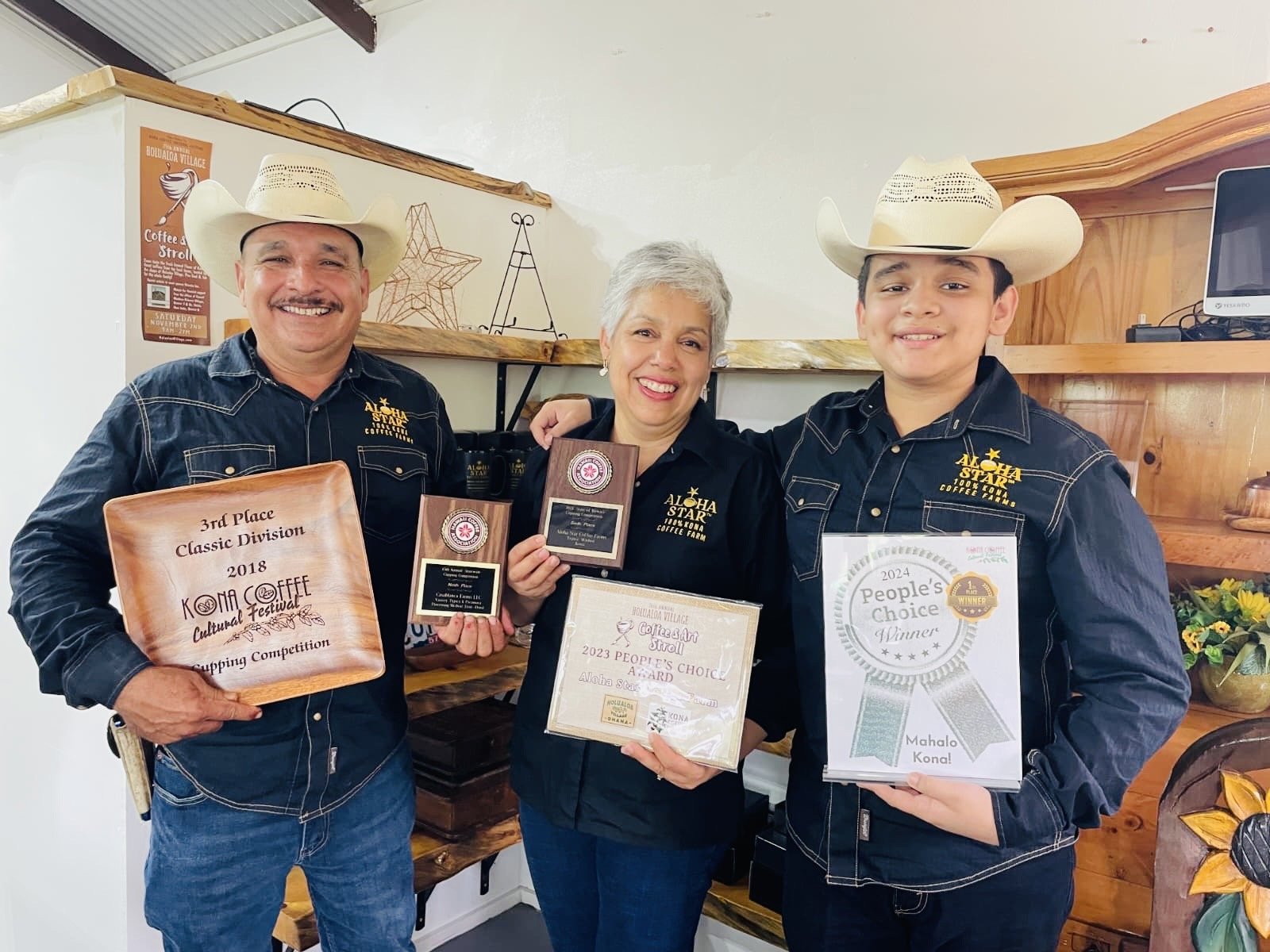 Three people smiling and holding awards at a coffee competition, wearing cowboy hats and black shirts with an Aloha Star Coffee Farm logo, standing in front of shelves with decorative items and flowers.