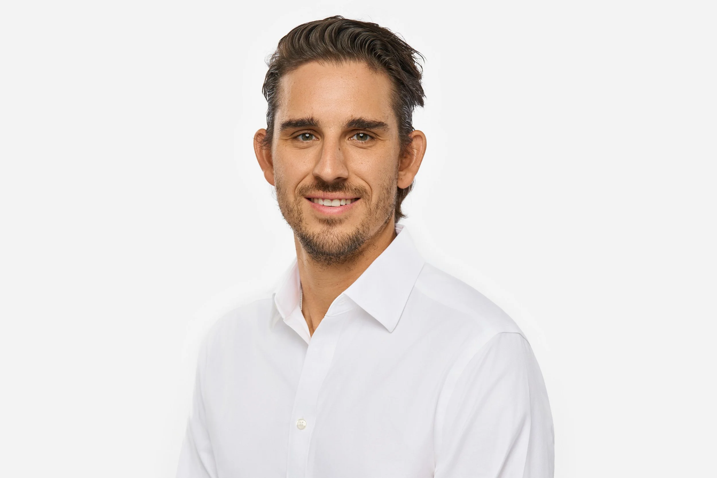 A professional headshot of a young man with brown hair, blue eyes, and light facial hair, wearing a white collared shirt, smiling against a white background.