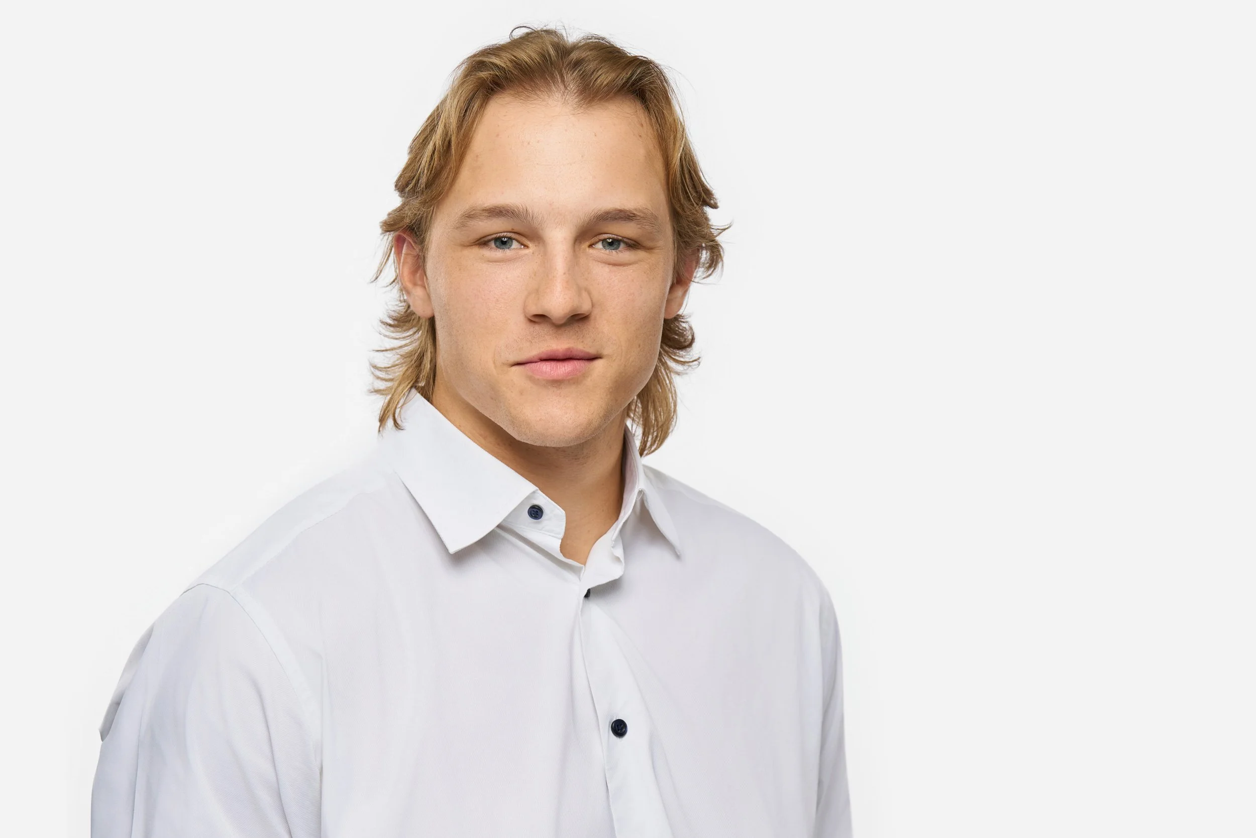 A professional headshot of a young man with shoulder-length blond hair, blue eyes, wearing a white dress shirt, standing against a plain white background.
