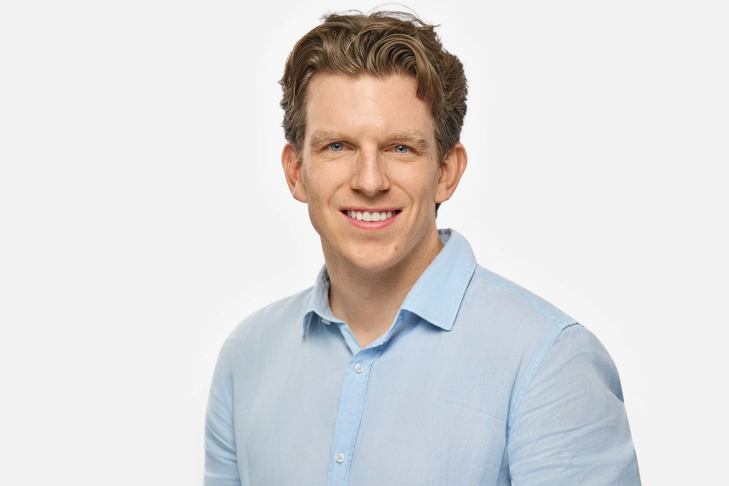 Professional Headshot of a man with brown hair, blue eyes, and a light blue collared shirt smiling against a plain white background.