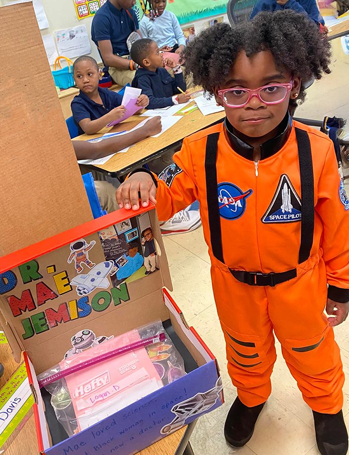 A young student in an astronaut costume next to her project about Dr. Mae Jemison