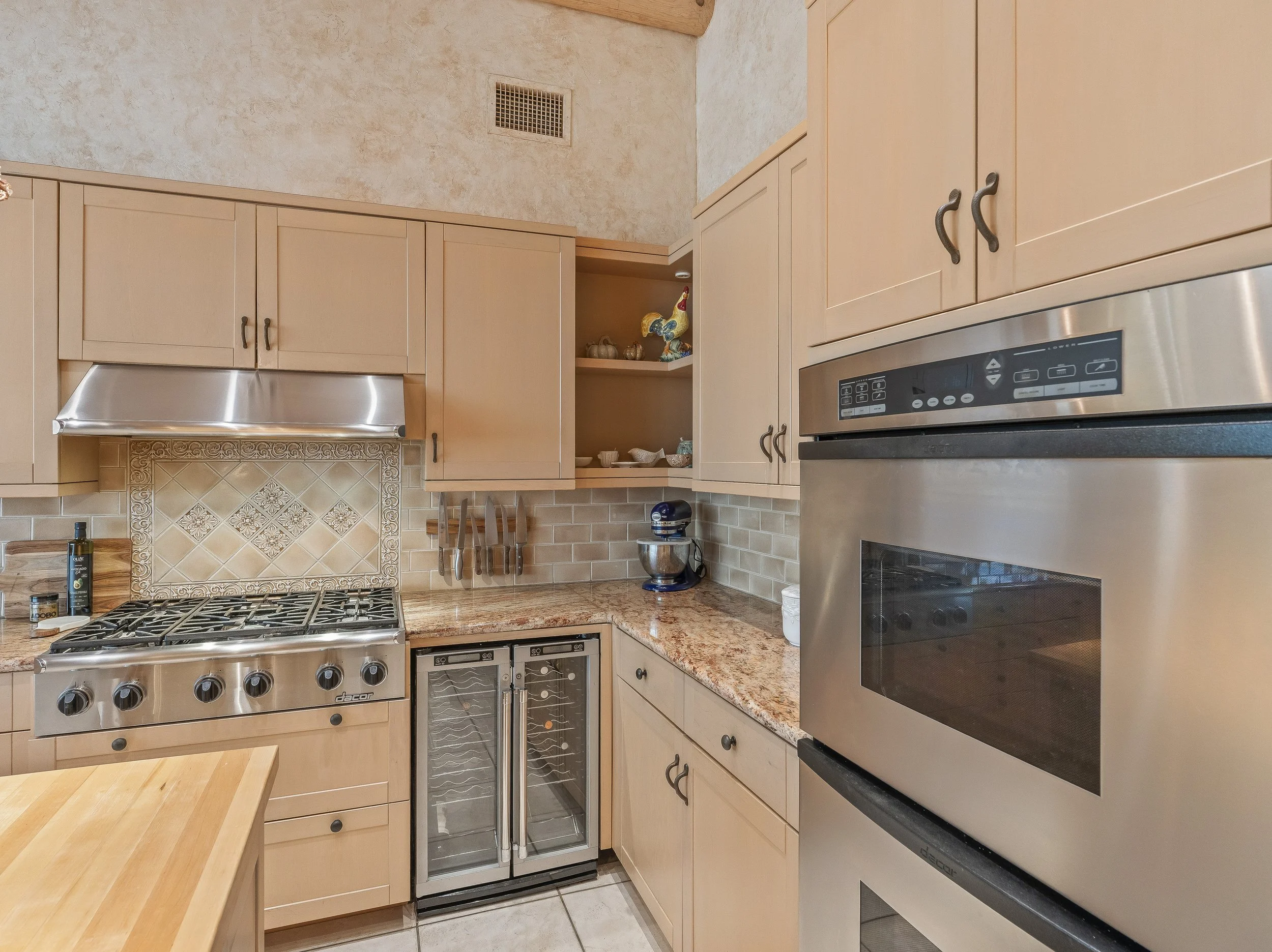 Spacious chef's kitchen with slab granite island, stainless steel gas cooktop, custom light wood cabinetry, and exposed viga ceiling beams in a luxury Tucson home.
