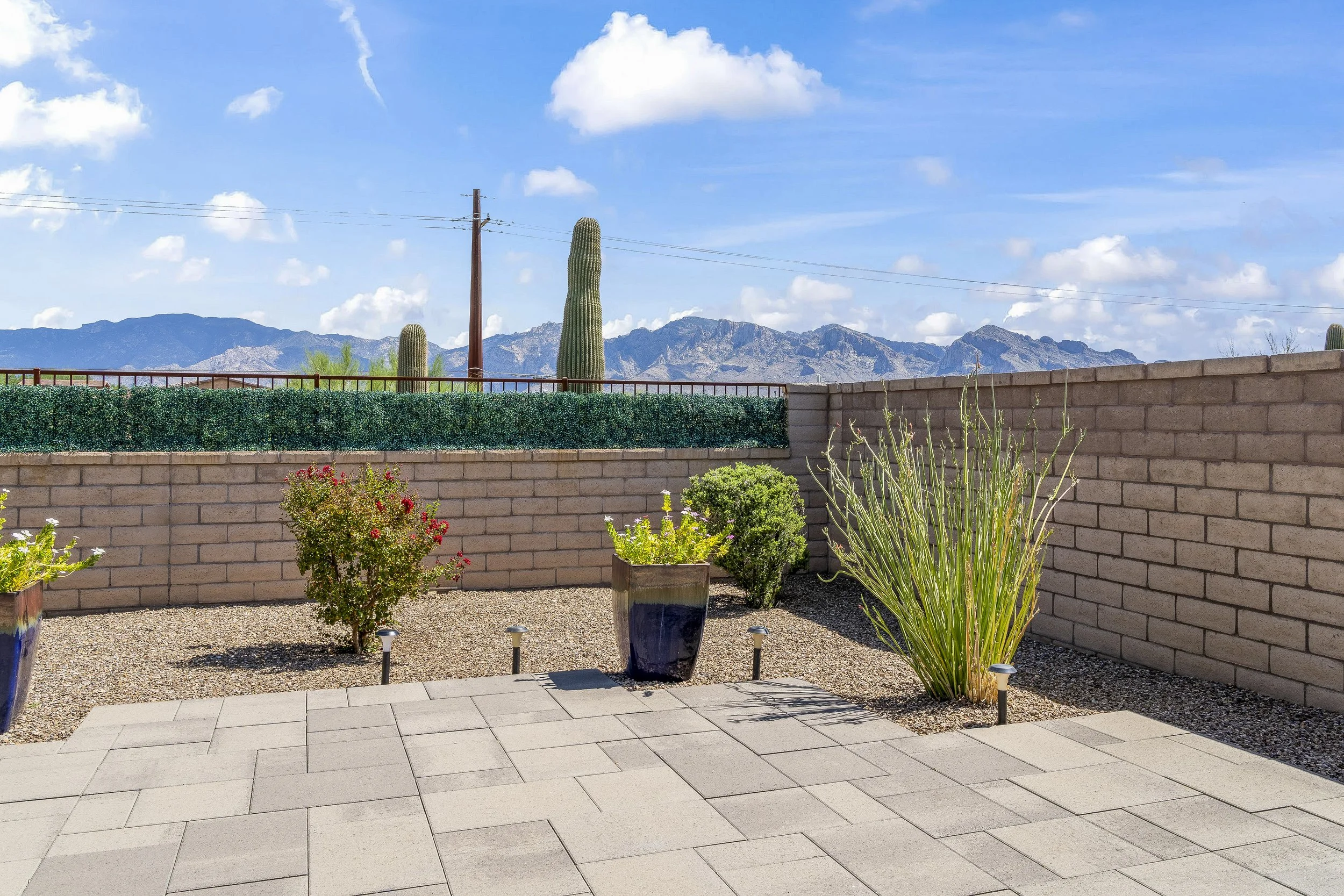 A sunny outdoor living space featuring a grey paver patio and low-maintenance desert landscaping with potted plants. The yard is enclosed by a privacy block wall, offering unobstructed views of distant mountains under a bright blue sky.