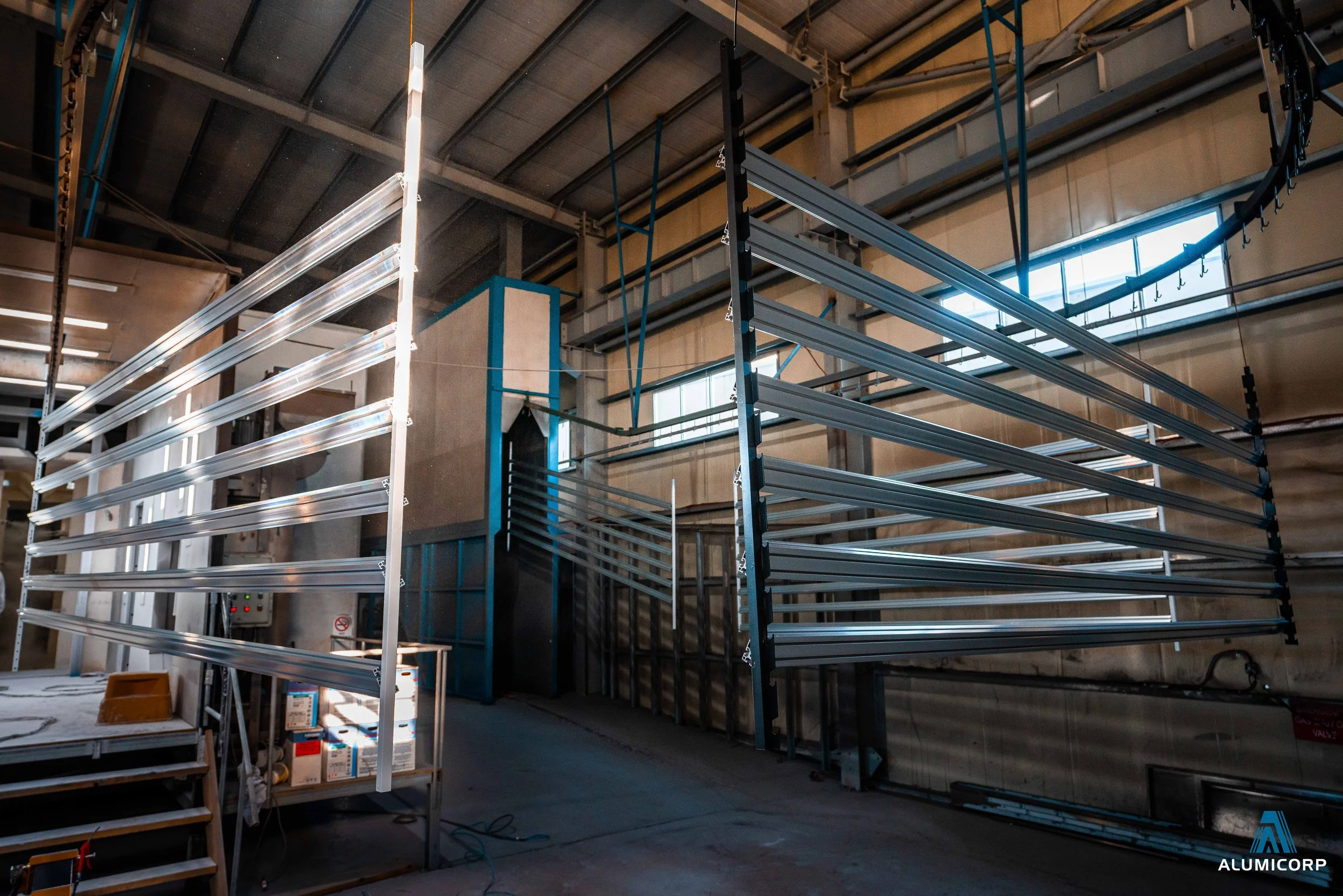 An industrial warehouse or factory interior with large aluminum racks and shelving units, some of which are empty, and a small work table with boxes and tools, illuminated by natural light from large windows.