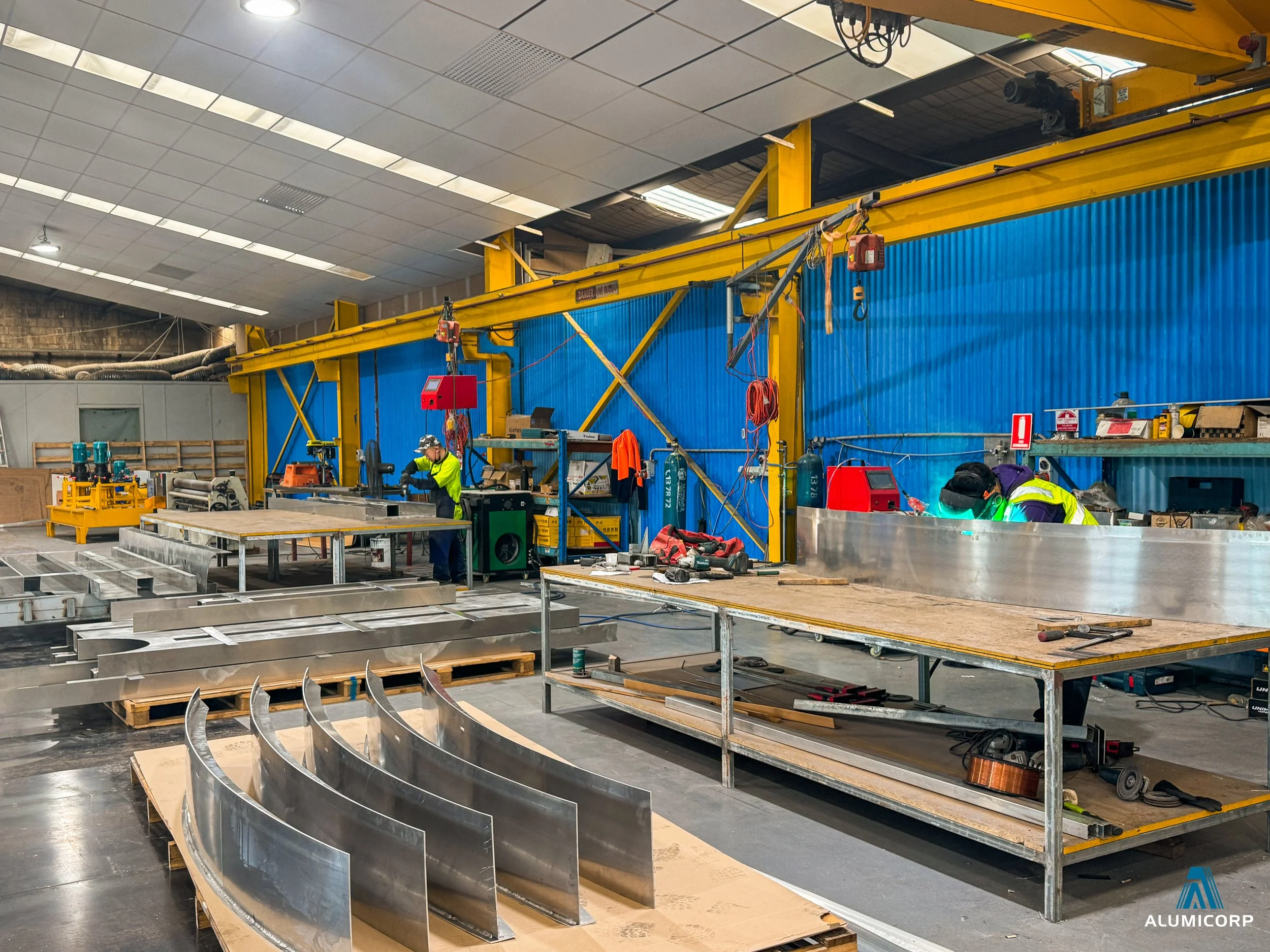 Industrial workshop with workers welding and assembling metal structures, tools and equipment on workbenches, large curved metal pieces in the foreground, and a yellow lifting crane overhead.