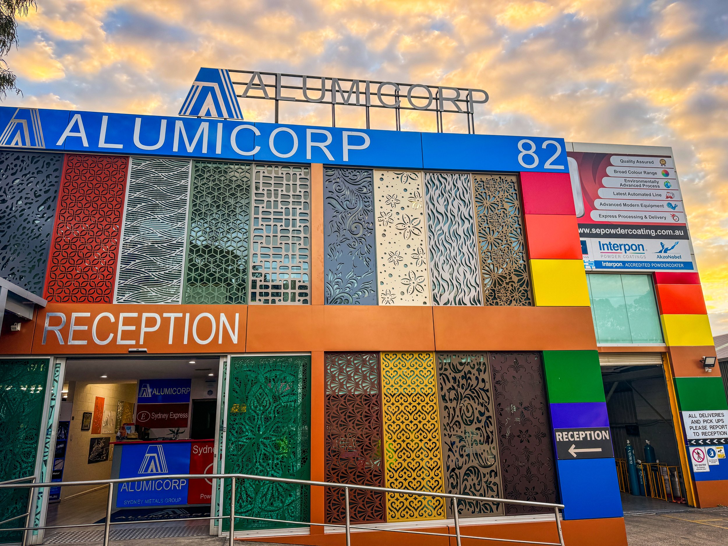 Colorful building exterior with decorative panels, signage for ALUMICORP, and a reception area with an open door, under a sunset sky.