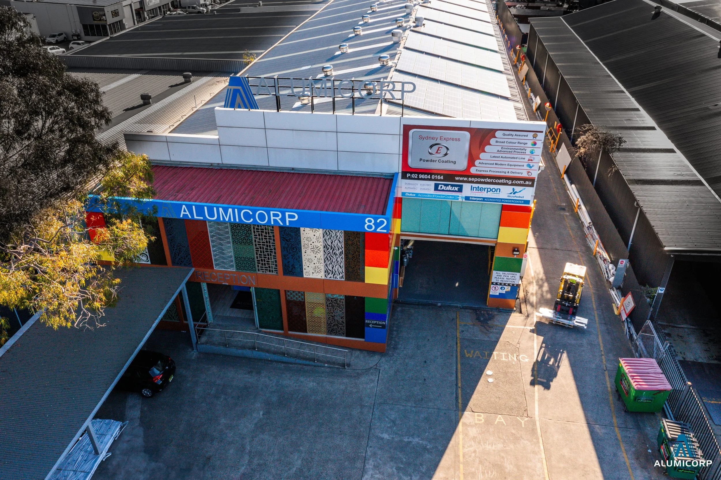An aerial view of a commercial building with colorful exterior walls and signage reading 'ALUMICORP'. The building has a parking area and a pathway marked 'WAITING BAY'. There are adjacent industrial warehouses and a forklift in the parking lot.