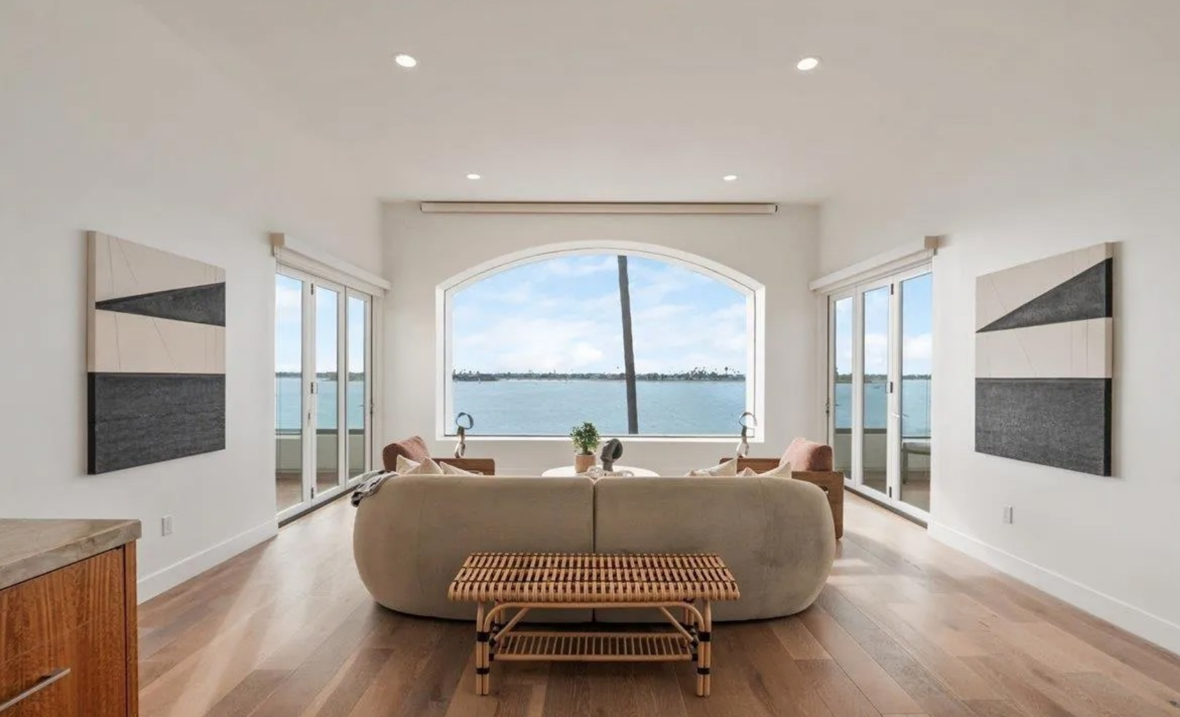 Living room with large window overlooking water, beige sofa, wooden chairs, artwork on white walls, and wooden floor.