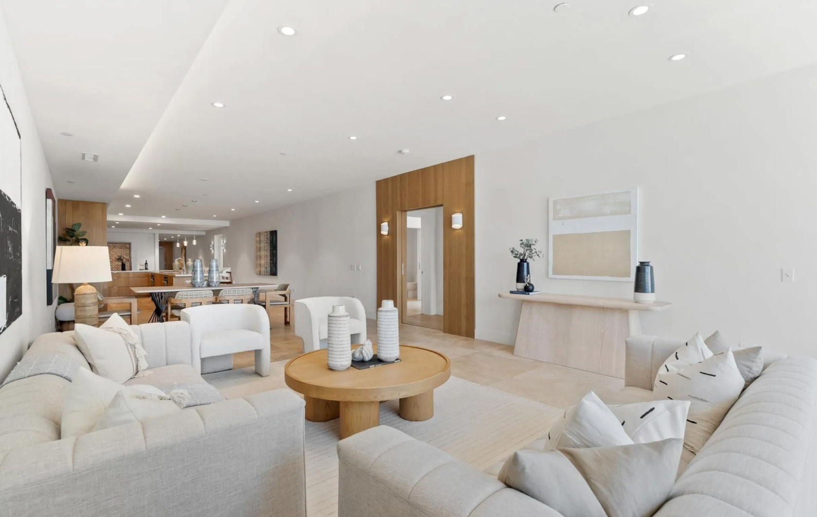 Modern living room with white sofas, wooden coffee table, and decorative vases, with dining area and hallway in the background.