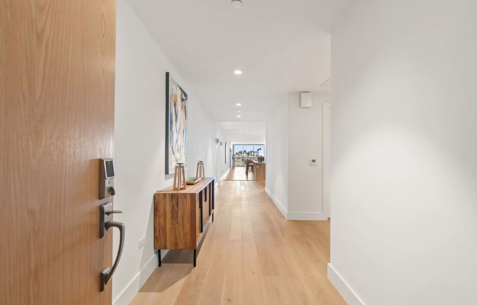 A bright, modern hallway with light wood flooring, white walls, and a wooden console table on the left side decorated with art and small stools. At the end, large windows reveal a view of palm trees and an outdoor area.