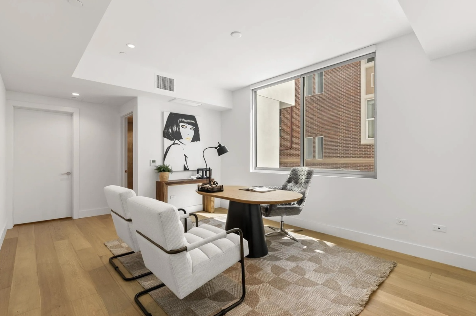 Modern office with white walls, wooden floors, a large window, a round wooden desk, an upholstered office chair, two white guest chairs, a black desk lamp, a small wooden table with a potted plant, a large black-and-white artwork of a woman with a bo