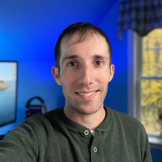 A young man taking a selfie in a room with a blue wall, window with curtains, and a computer monitor in the background.