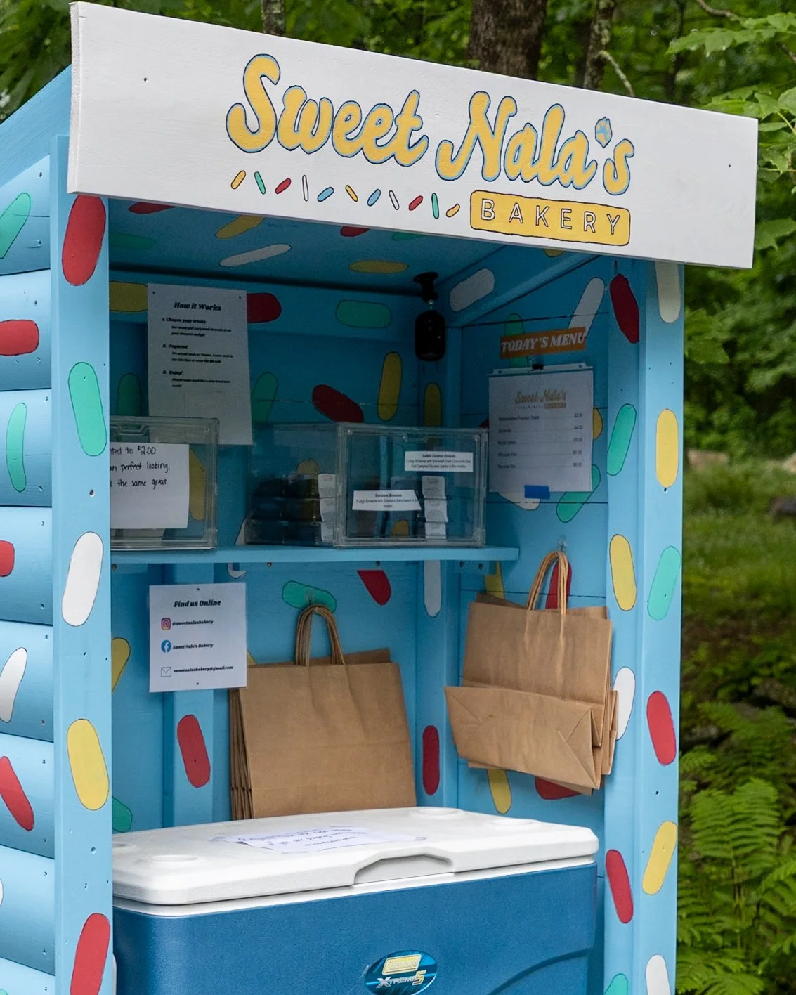 A colorful ice cream stand named 'Sweet Nala's Bakery' with a light blue exterior decorated with multicolored sprinkles. It has a sign on the top with the shop's name and a small menu board, paper bags hanging on the side, and a white cooler at the bottom. There are trees and foliage in the background.