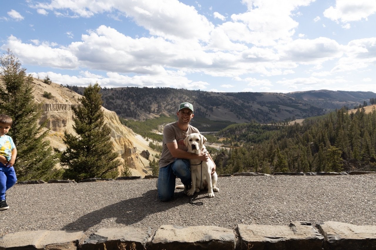 A man, a dog, and a young boy at a scenic mountain overlook with trees, hills, and clouds in the sky.