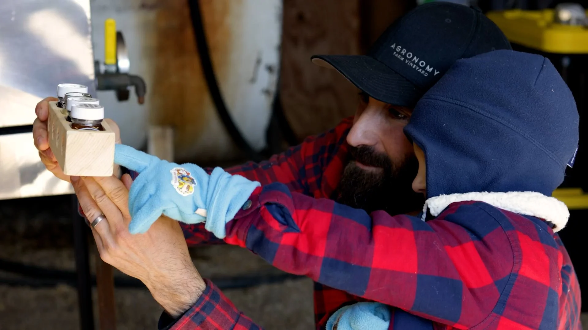 A man and a child examining or holding a wooden block with glass jars inside it, both wearing hats and gloves, in a workshop or outdoor setting.