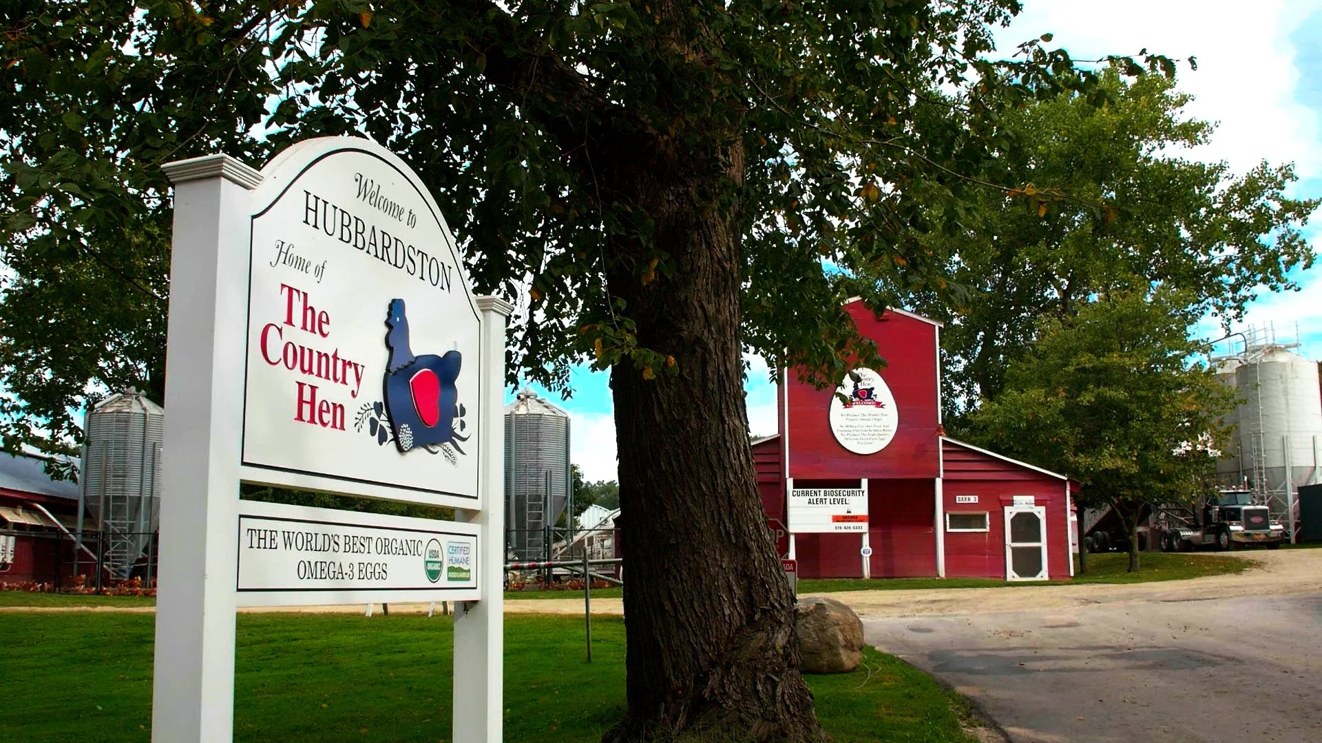 Sign welcoming visitors to Hubbardston, featuring a blue hen graphic, and an image of a red barn with farm silos in the background, surrounded by trees and a gravel driveway.