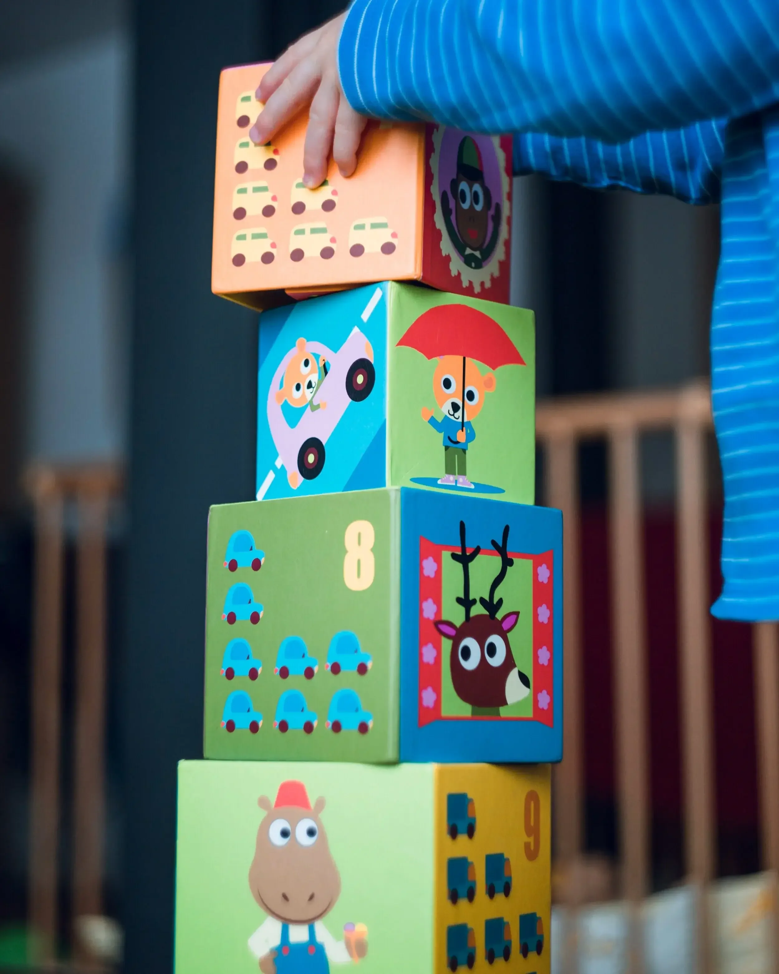 A child’s hand stacking colorful blocks with playful animal and number designs. The blocks feature images such as a deer, a cow, cars, and numbers eight and nine.