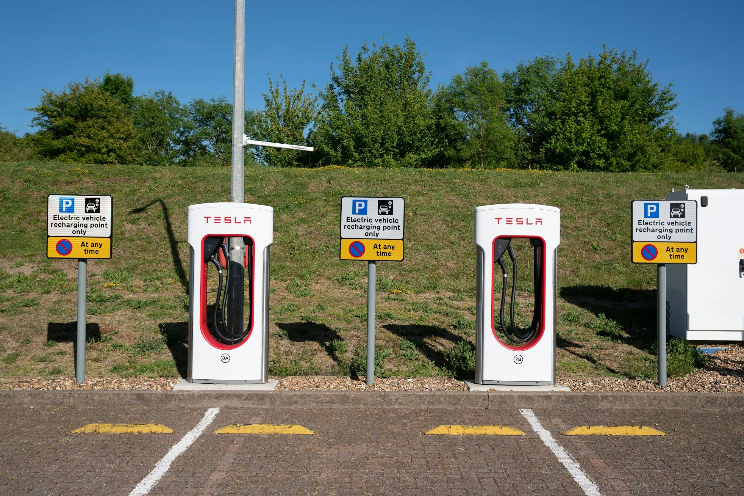 Four electric vehicle charging stations with signs indicating charging only for electric vehicles, located in a parking lot with painted parking spots and a grassy area with trees in the background.