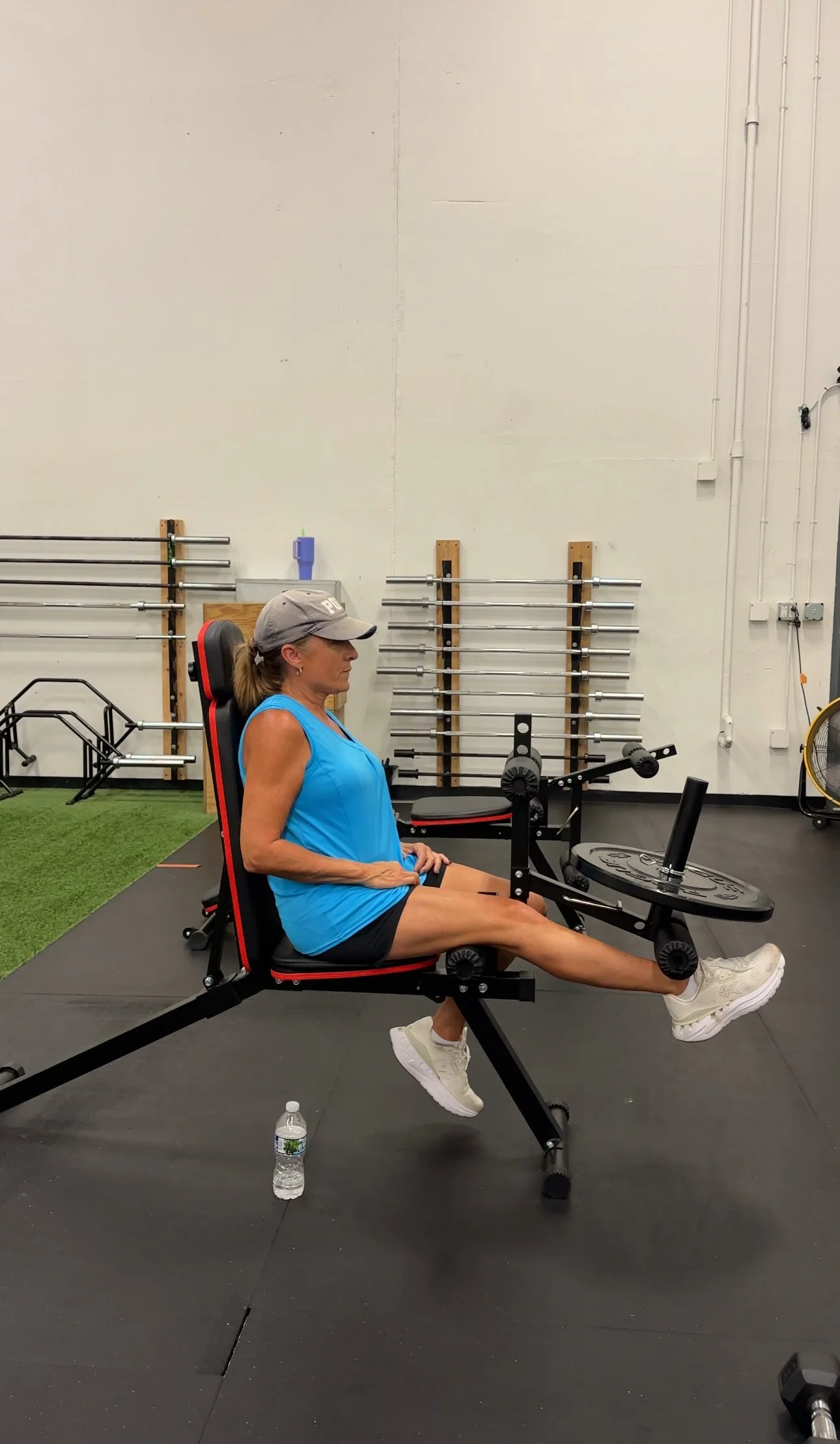 Woman exercising on a seated leg curl machine in a gym, wearing a gray cap, blue tank top, black shorts, and white sneakers.