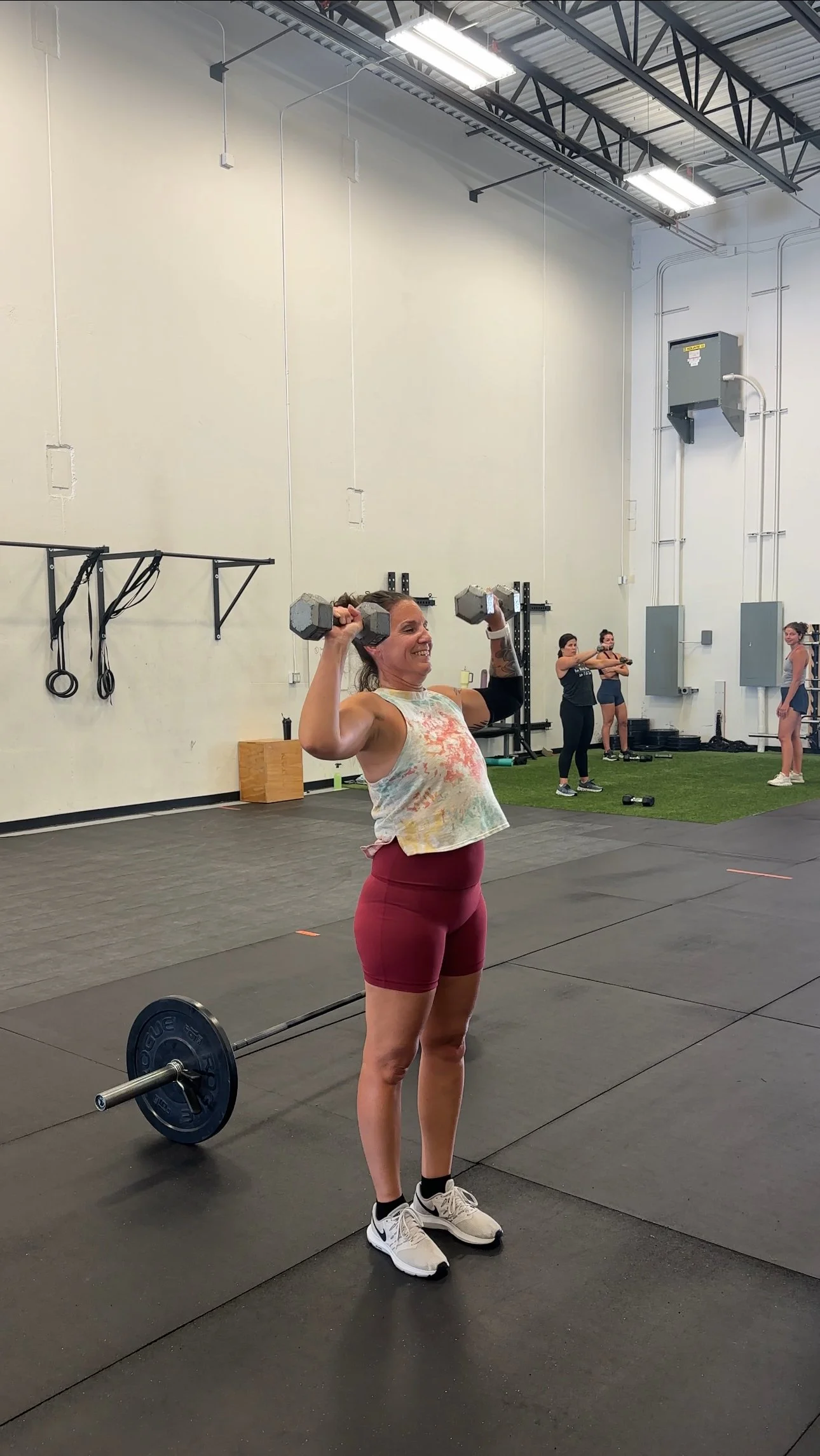 A woman lifting dumbbells during a workout session at a gym. She is smiling, wearing a sleeveless tie-dye shirt, red shorts, and athletic shoes. In the background, there are other women exercising.