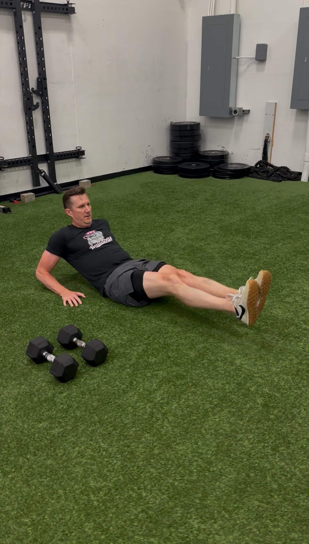 A man performing a seated abdominal exercise on artificial turf in a gym, with a black T-shirt, gray shorts, and white sneakers, two dumbbells nearby, and workout equipment in the background.