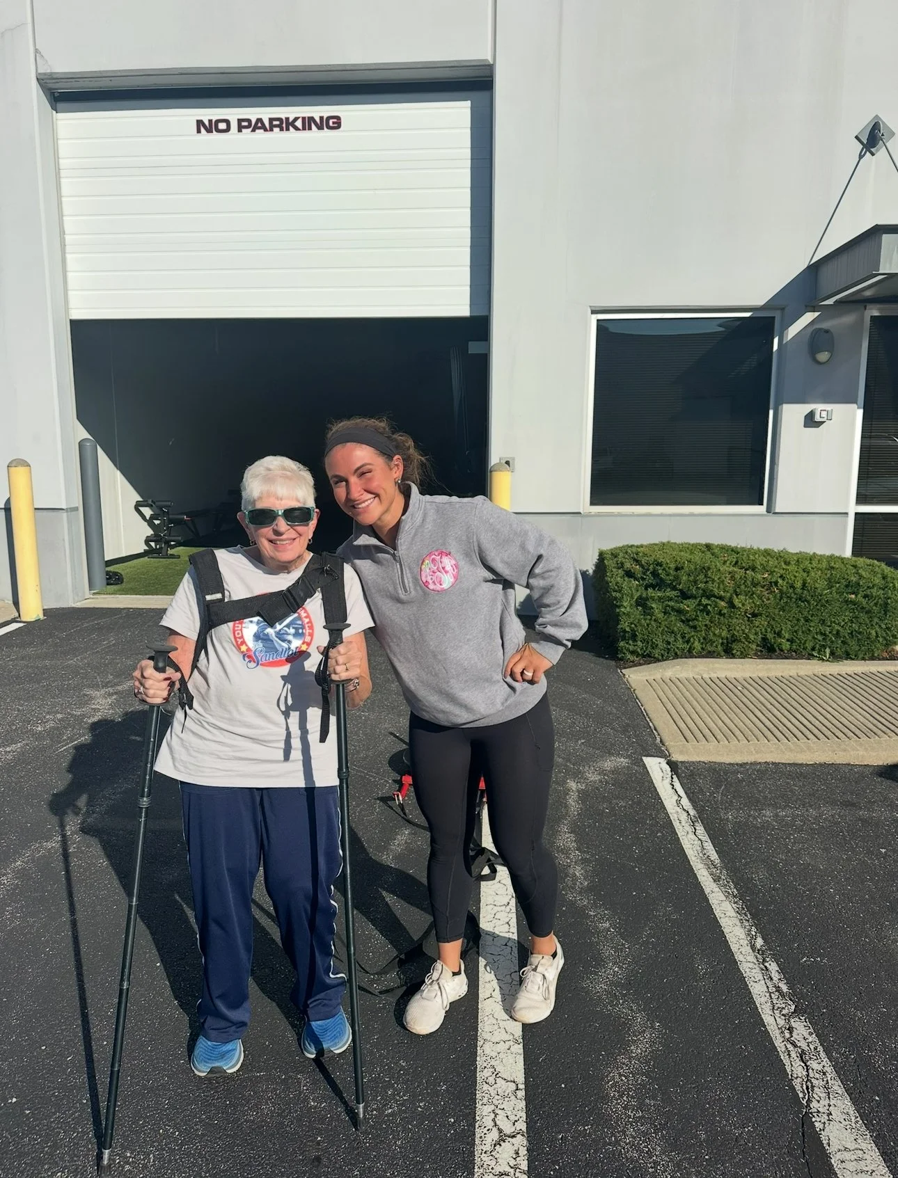 Two women standing outside in a parking lot, smiling, with a building and a garage door behind them. One woman is elderly, wearing sunglasses, a white T-shirt, and holding hiking poles. The other woman is younger, wearing a gray sweatshirt, black leggings, and white sneakers.