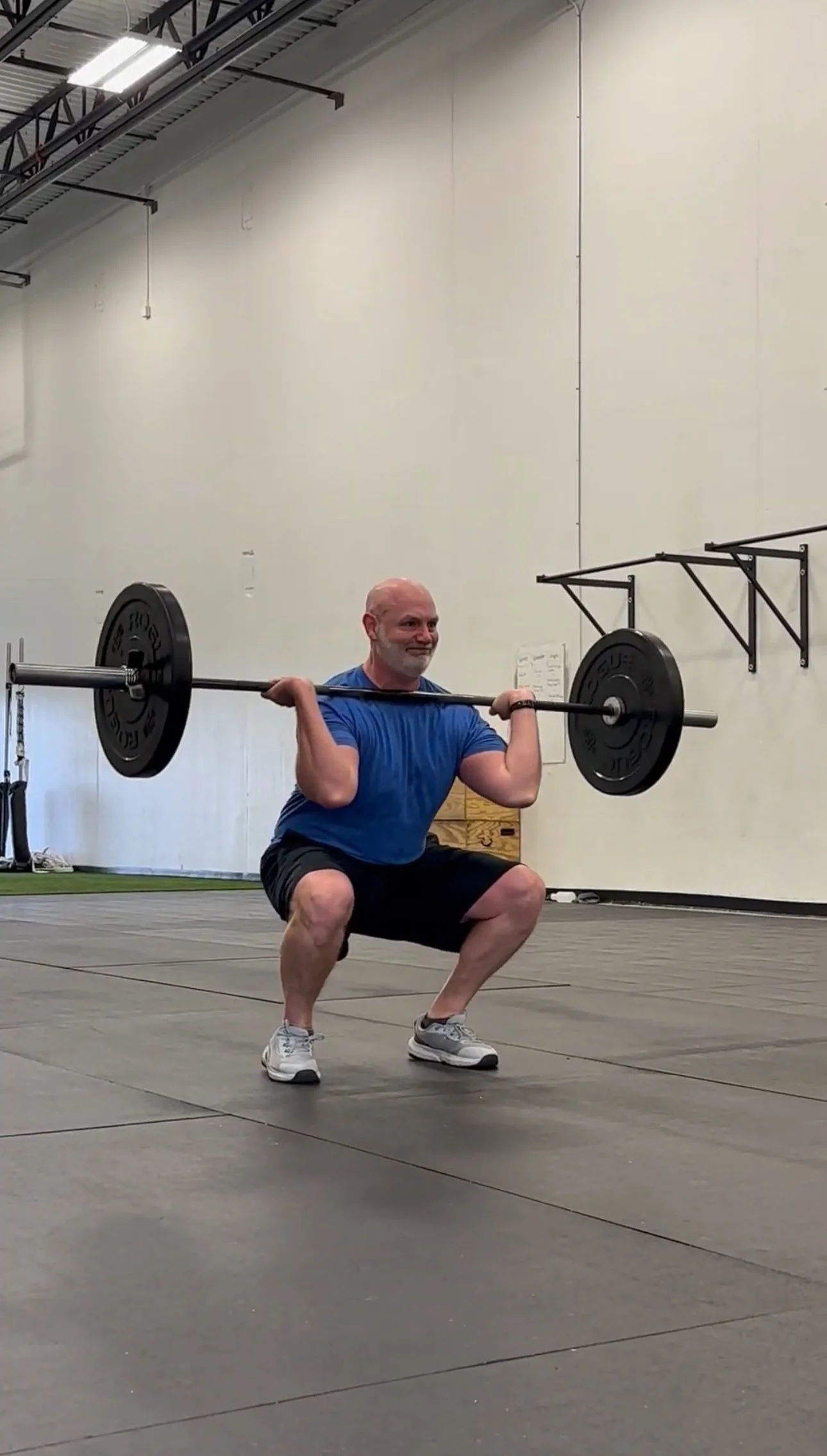 A man with a bald head and gray beard performing a barbell squat exercise in a gym with a plain wall background. He is wearing a blue t-shirt, black shorts, and gray sneakers.
