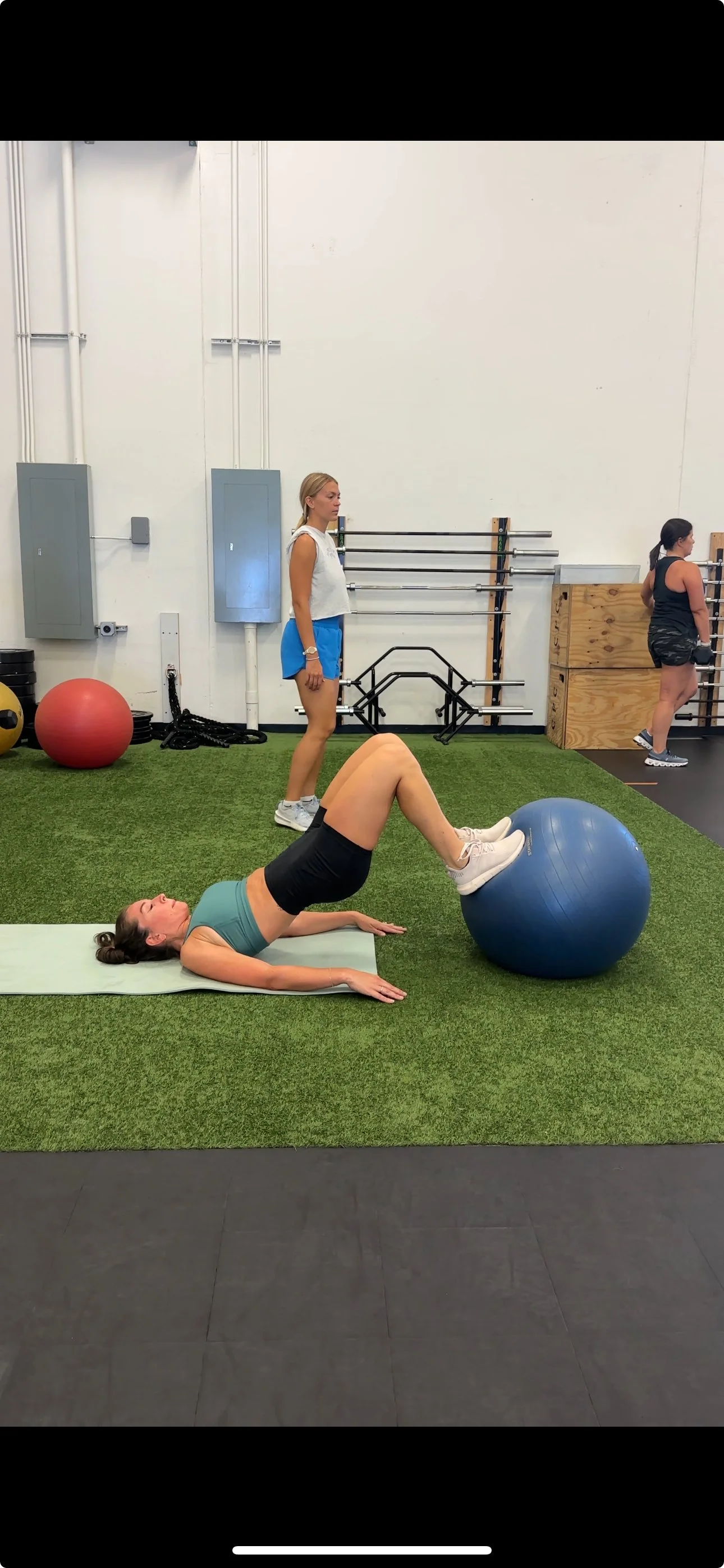Woman lying on exercise mat performing a glute bridge with a stability ball under her legs at gym, with two women standing nearby and gym equipment in the background.