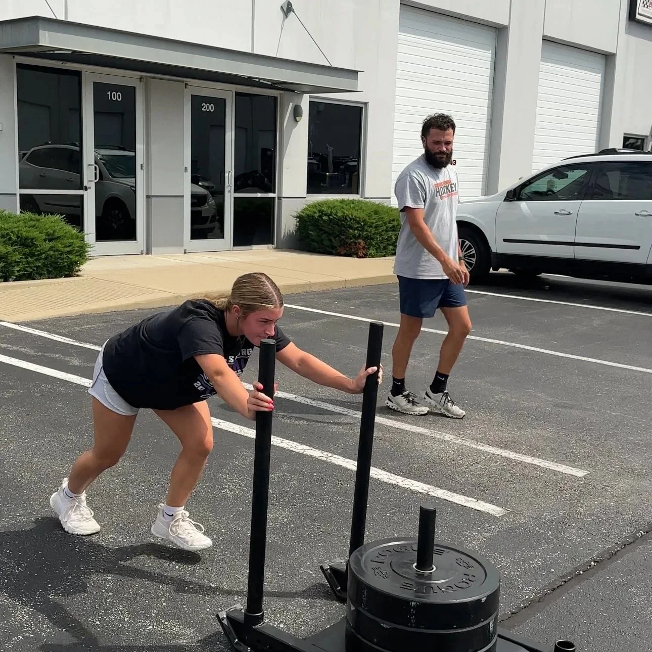 A woman with curly hair, glasses, hoop earrings, and layered necklaces, smiling in an indoor setting with a beige wall and window in the background.