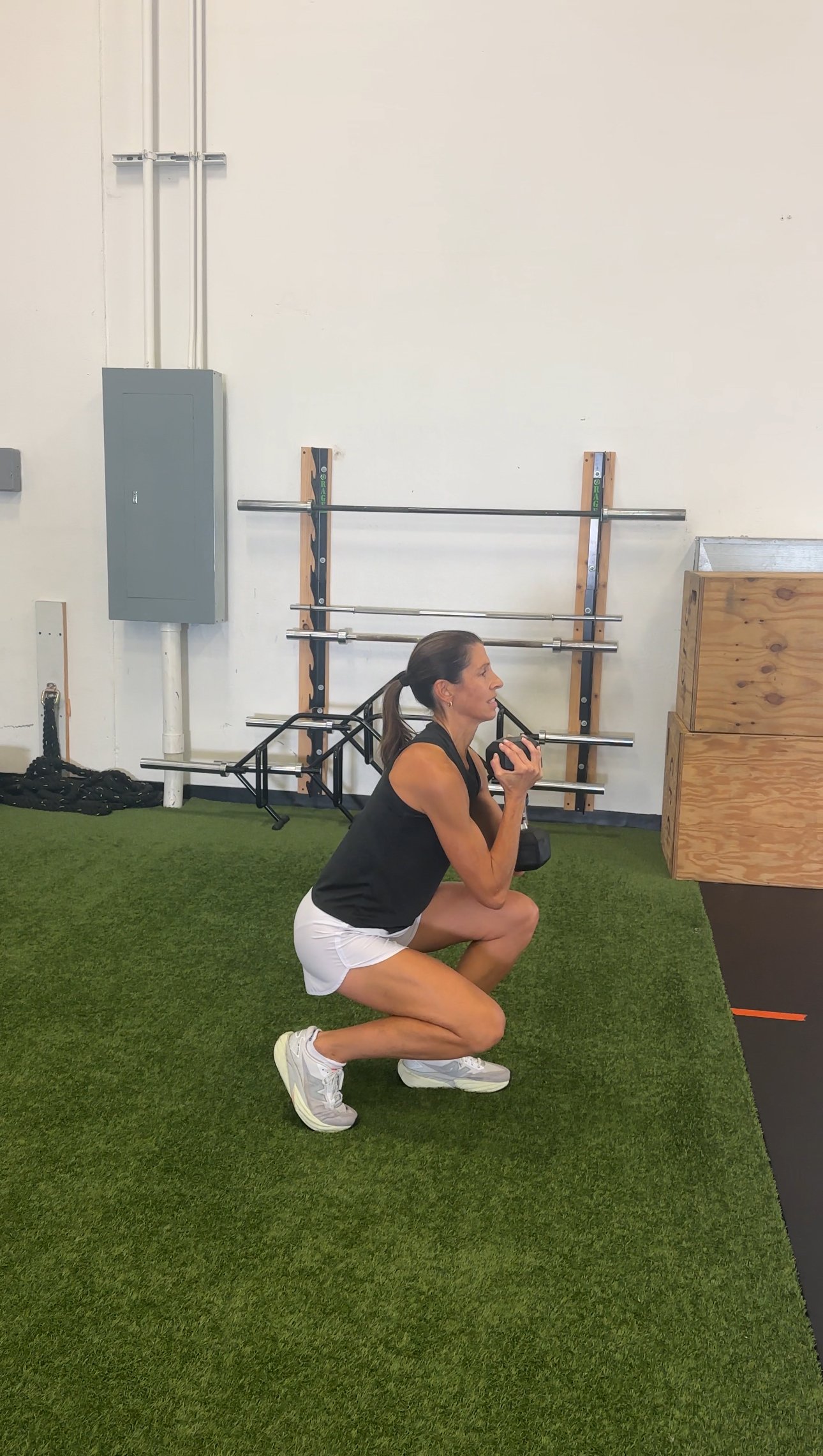 A woman Squatting on a gym floor, holding a kettlebell close to her chest, wearing a black tank top, white shorts, and athletic shoes, in a gym with workout equipment in the background.