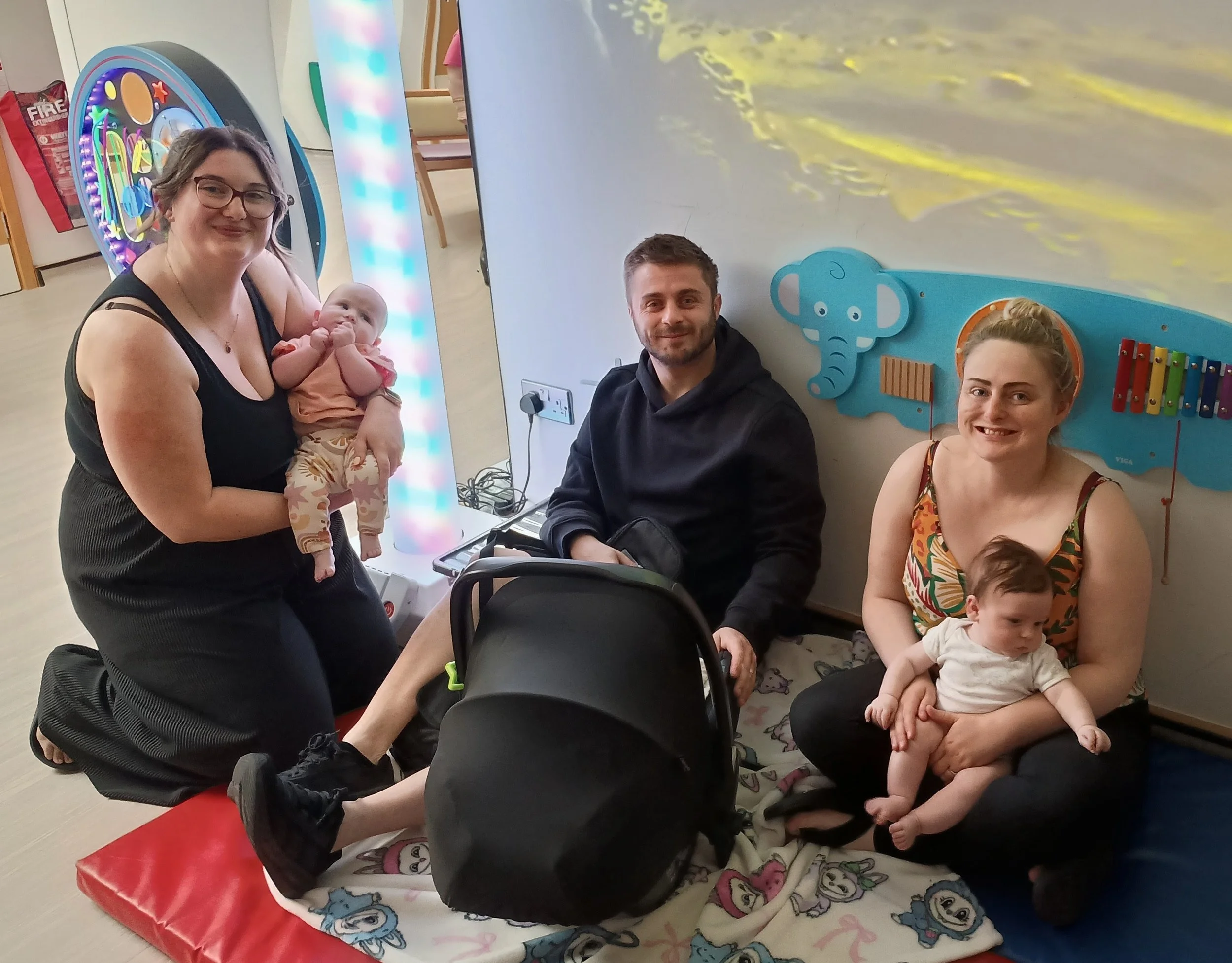 Three people sitting on the floor holding their babies in a colourful room with toys
