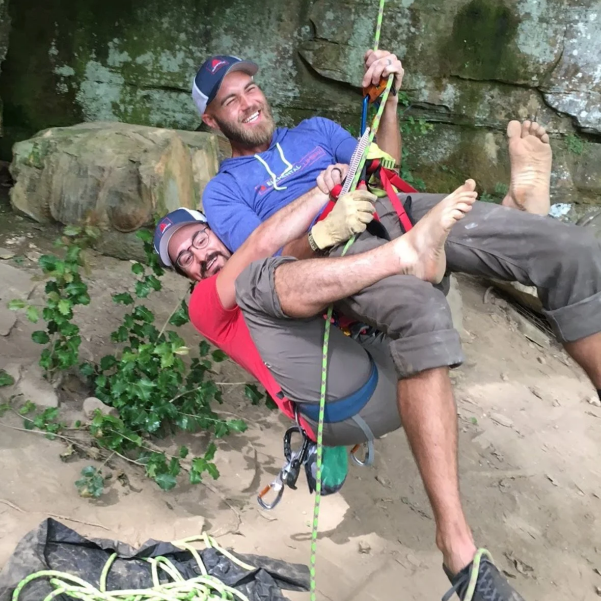 Two men, wearing climbing gear and helmets, crouch on a dirt ground in front of a rocky wall, smiling and posing playfully. One man is holding onto a climbing rope.