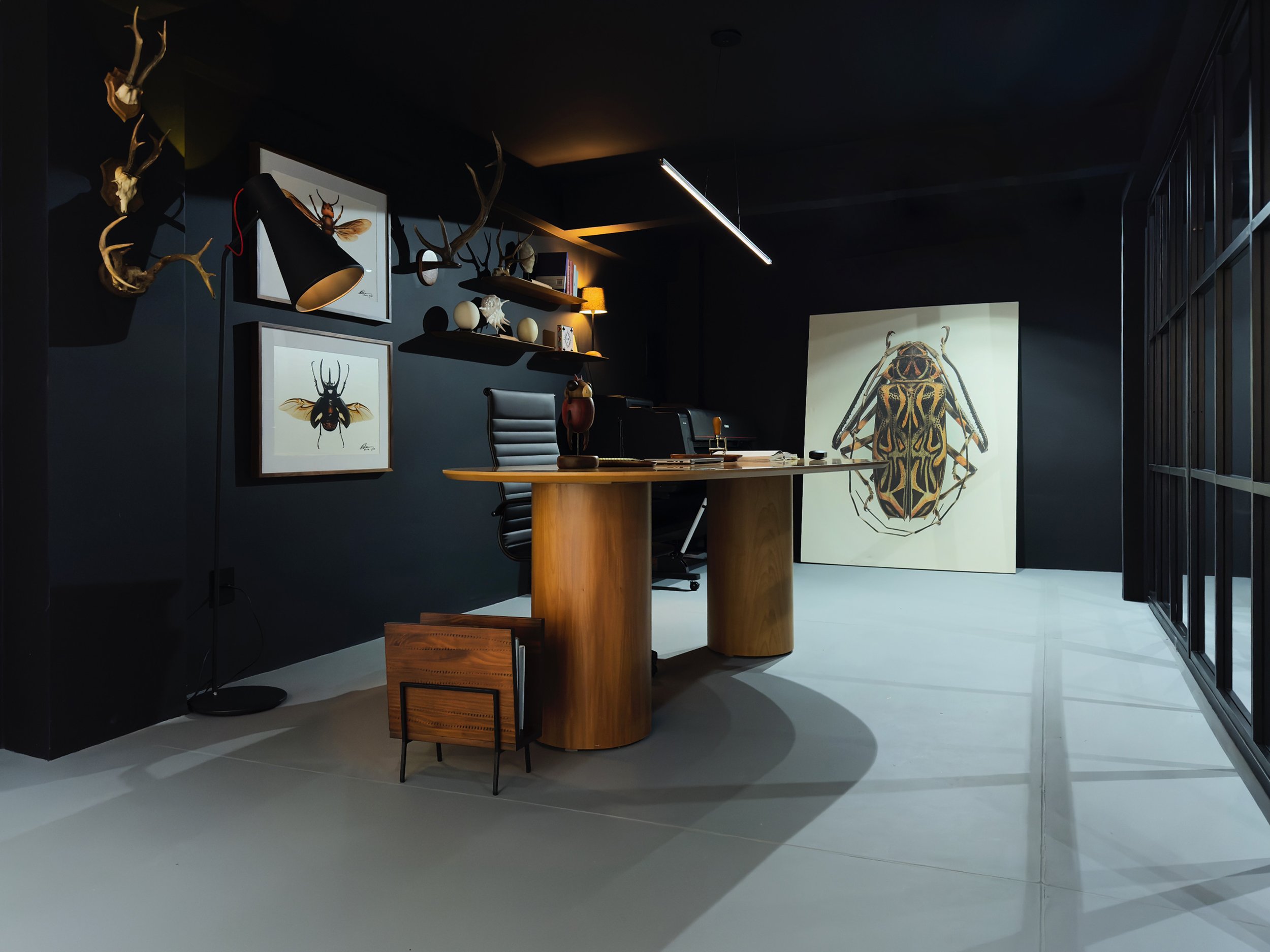 Sala de escritório decorada com tema de insetos e animais, incluindo quadros de insetos, chifres de cervo na parede, uma grande pintura de um inseto no chão, mesa de madeira com cadeira de escritório, prateleiras com objetos decorativos, luzes e uma 