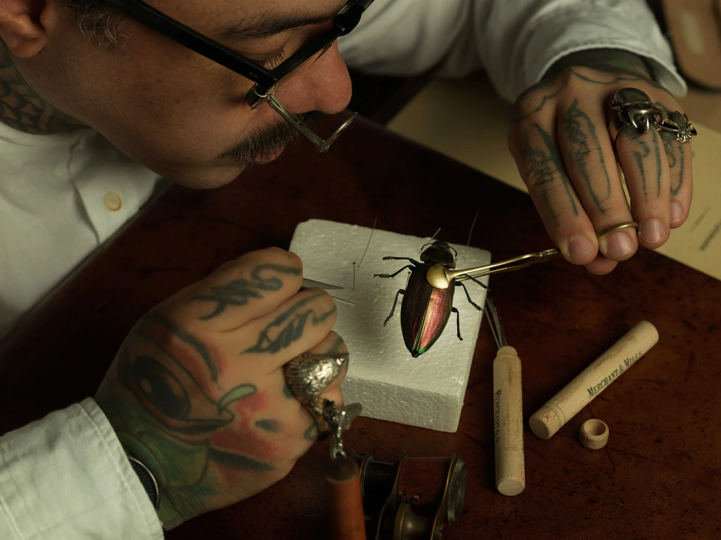 Pessoa tatuada trabalhando na montagem de uma joia, usando ferramentas e apoiada em uma mesa de madeira, com foco na joia de inseto.