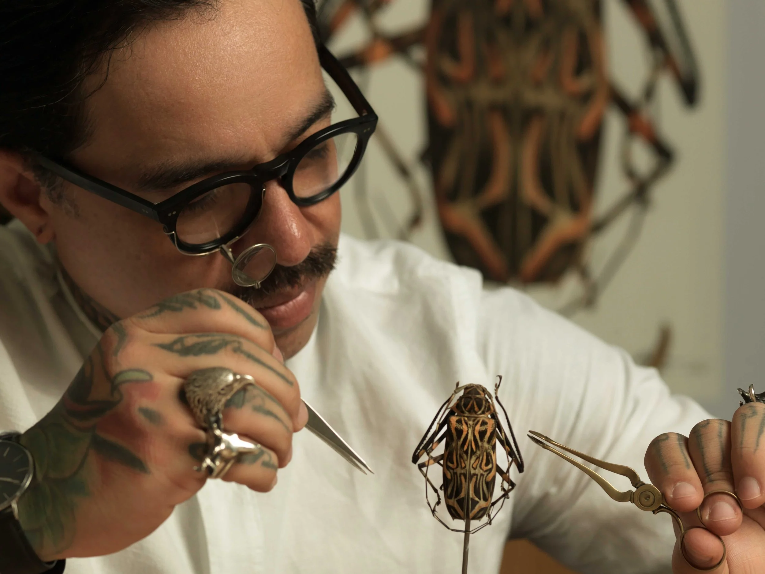 Homem tatuado, com óculos e piercing no nariz, estudando uma joia ou inseto ornamentado com ferramentas de precisão, enquanto olha atentamente para o objeto, com uma obra de arte tribal ao fundo na parede.