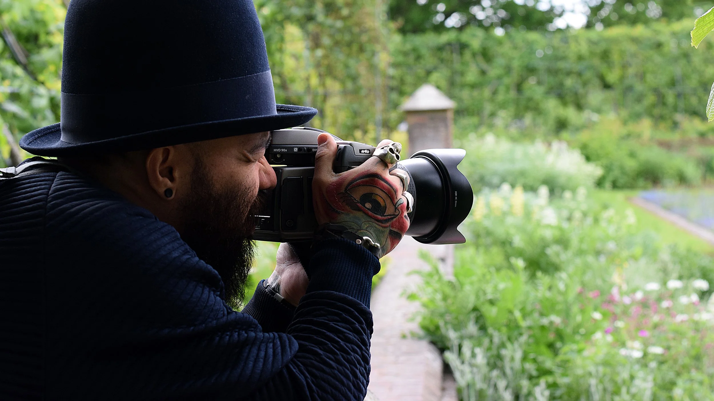 Homem com tatuagem na mão, usando chapéu preto, tirando uma foto com uma câmera DSLR em um ambiente ao ar livre com vegetação verde e um caminho de pedra.