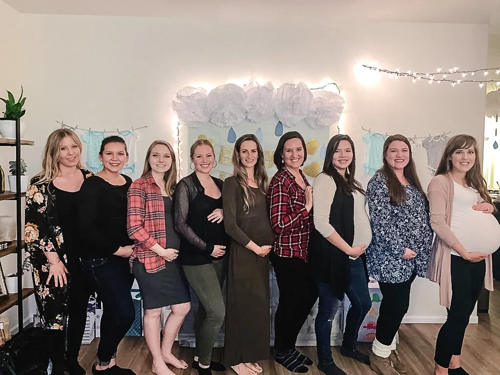 A group of pregnant women smiling and posing together with Sierra Madre Midwifery. The setting is decorated with soft lighting and baby-themed decorations on the wall. The women are standing closely side by side indoors. Featuring Kattie Jones.