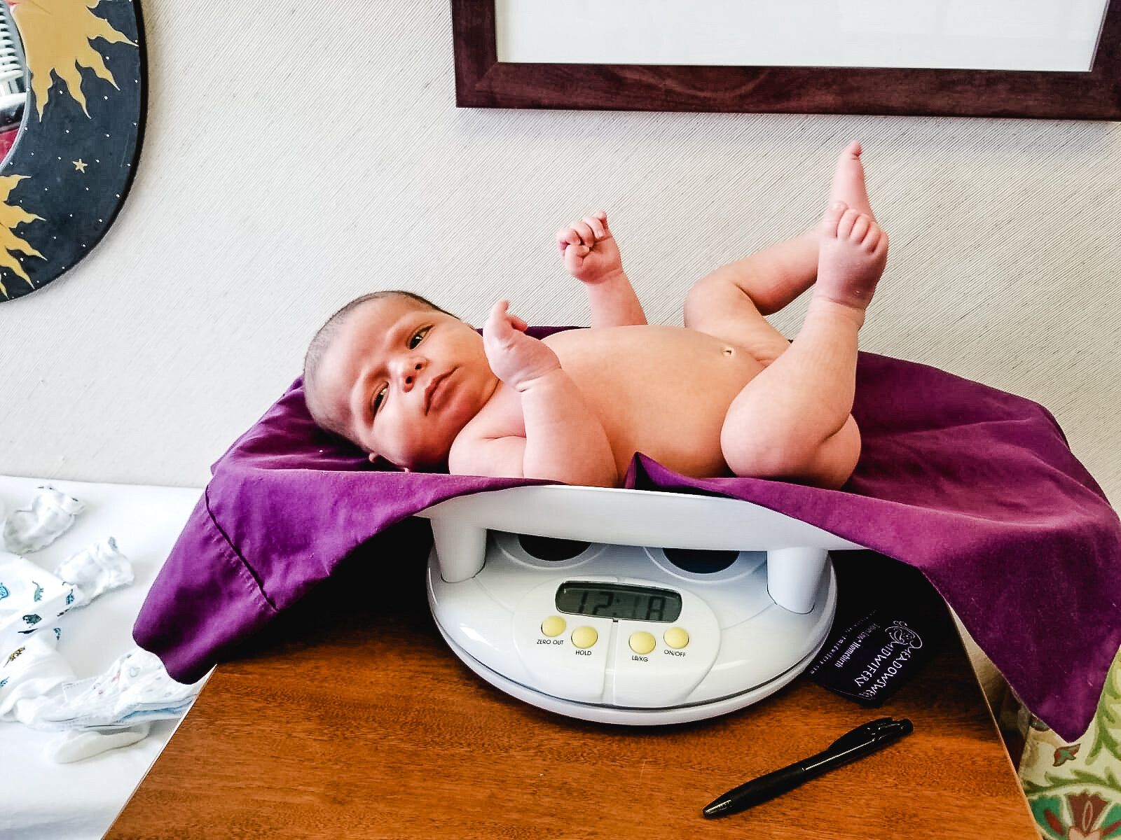 A newborn baby lying on a digital scale, covered with a purple cloth, in a room with decorative elements on the wall. The baby was delivered by sierra madre midwifery or Kattie jones, a midwife who offers home birth options in denver co