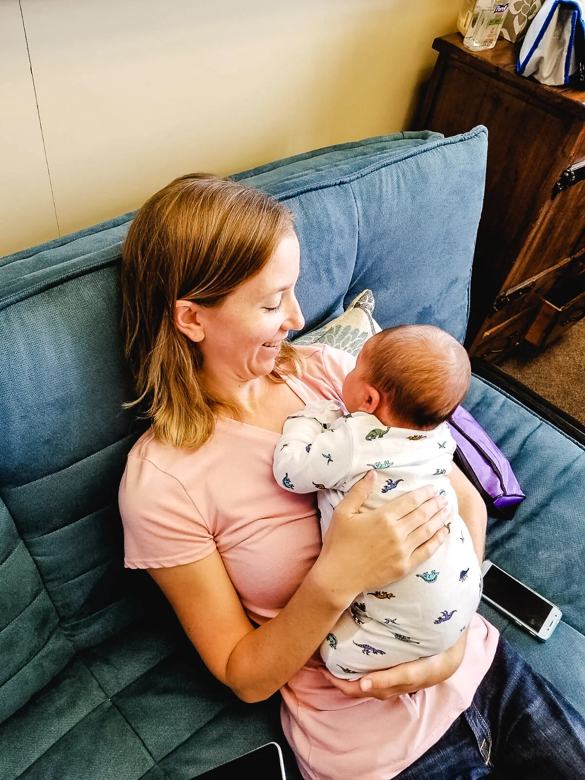 A woman in a pink shirt is sitting on a blue couch, smiling and holding a baby dressed in a dinosaur-patterned onesie. Two smartphones are nearby on the couch. Happy after working with sierra madre midwifery in denver colorado
