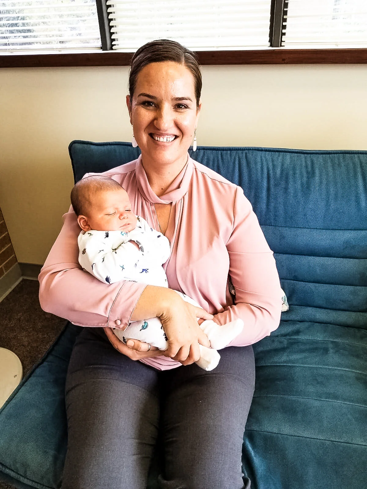 Kattie Jones sitting on a blue sofa holding a sleeping baby in her arms, wearing a pink blouse and smiling.