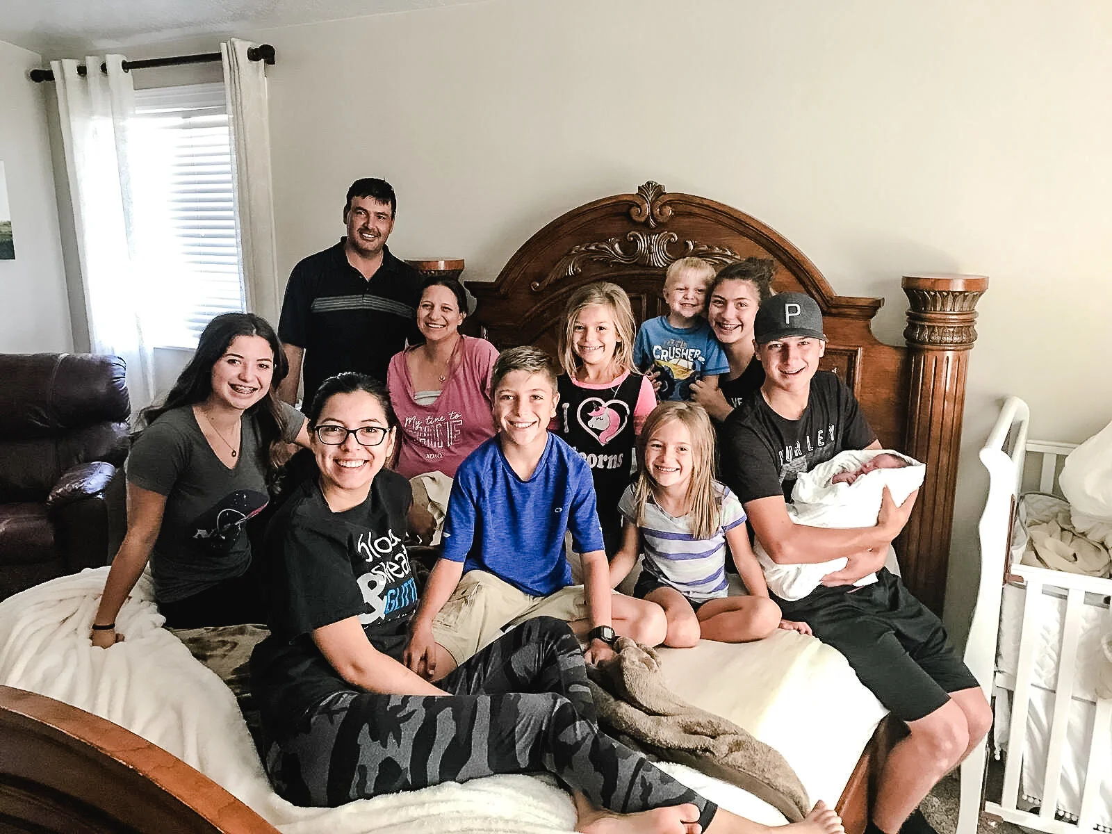 Group of people smiling in a bedroom with a wooden bedframe, including a newborn baby being held by a person seated at the edge of the bed. They are previous clients of sierra madre midwifery.