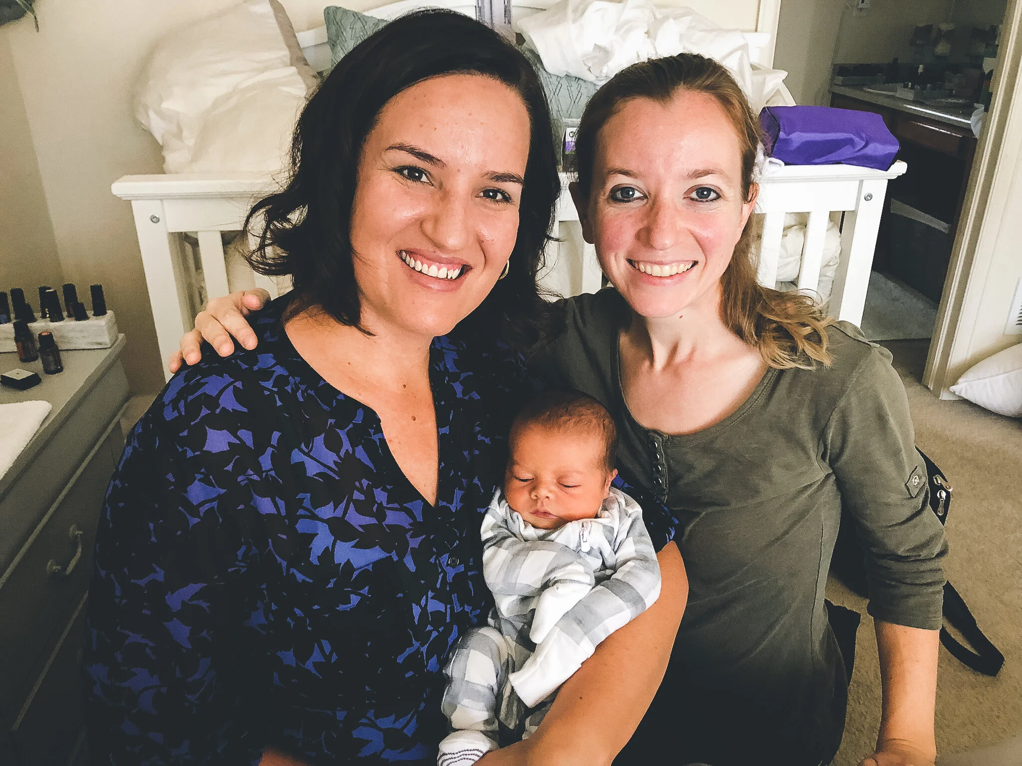 Two women smiling while posing with a newborn baby in a cozy room with baby items and essential oils in the background. One of them is kattie jones, a homebirth midwife that offers natural birth plans in denver co and surrounding areas.