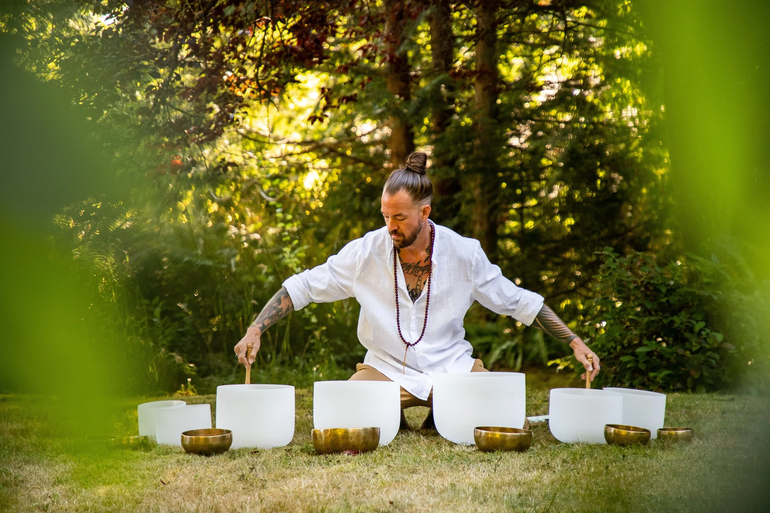 Man with tattoos in white shirt playing singing bowls outdoors on grassy field surrounded by trees.